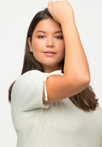 Top bianco a maniche corte con polsini legati, caratterizzato da una consistenza morbida e una vestibilità rilassata. Il modello ha capelli lunghi e scuri e un'espressione neutra.