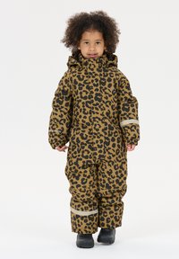 Barn med krøllet hår iført en snedragt i brunt og sort leopardprint med reflekterende striber og sorte støvler, står på en hvid baggrund.