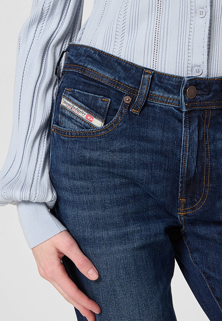 Jeans in denim blu scuro con vestibilità slim, caratterizzati da cuciture arancioni e un'etichetta del marchio visibile sulla tasca anteriore. Tessuto texturizzato e liscio.