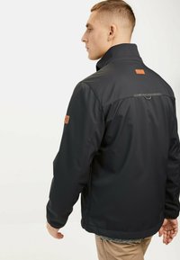 Veste synthétique noire avec un col monté, une étiquette en cuir marron au dos et un détail en boucle. Présente une finition lisse et texturée.