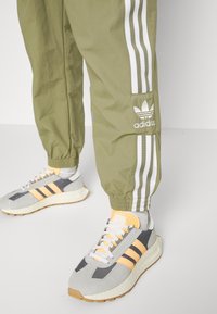 Ben som bär olivgröna Adidas träningsbyxor med vita ränder och grå, persiko- och vita Adidas sneakers på ett ljusgrått golv.
