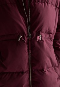 Primo piano di una giacca a vento borgogna con una zip argentata e due toggles per cordoncino vicino all'area del petto.