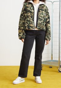 Miękka, zielono-beżowa kurtka zapinana na zamek z czarnymi wykończeniami, połączona z czarnymi dżinsami i białymi trampkami z zielonymi akcentami.