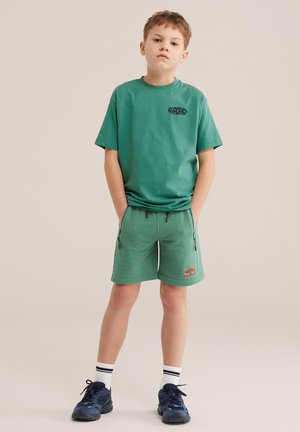 Ragazzo in piedi con le mani in tasca, indossa una maglietta verde, pantaloncini, calzini bianchi e scarpe da ginnastica blu scuro su uno sfondo neutro.