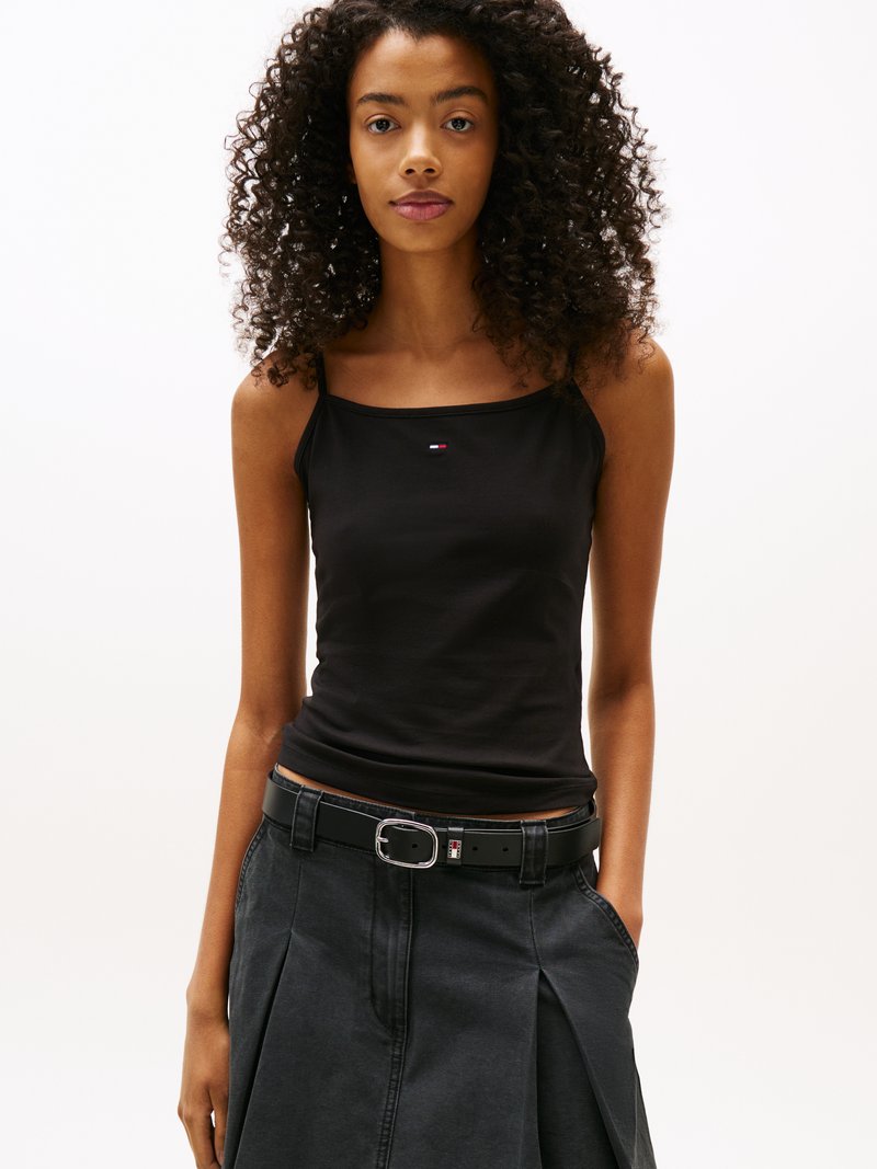 Black tank top with thin straps, featuring a small logo patch. Paired with a high-waisted, pleated black skirt and a black belt with a silver buckle.
