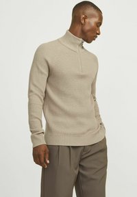 Beige geribbelde gebreide trui met een hoge kraag en een halve ritssluiting, voorzien van lange mouwen en een fitted ontwerp. Textuurdetails van de stof zijn zichtbaar.