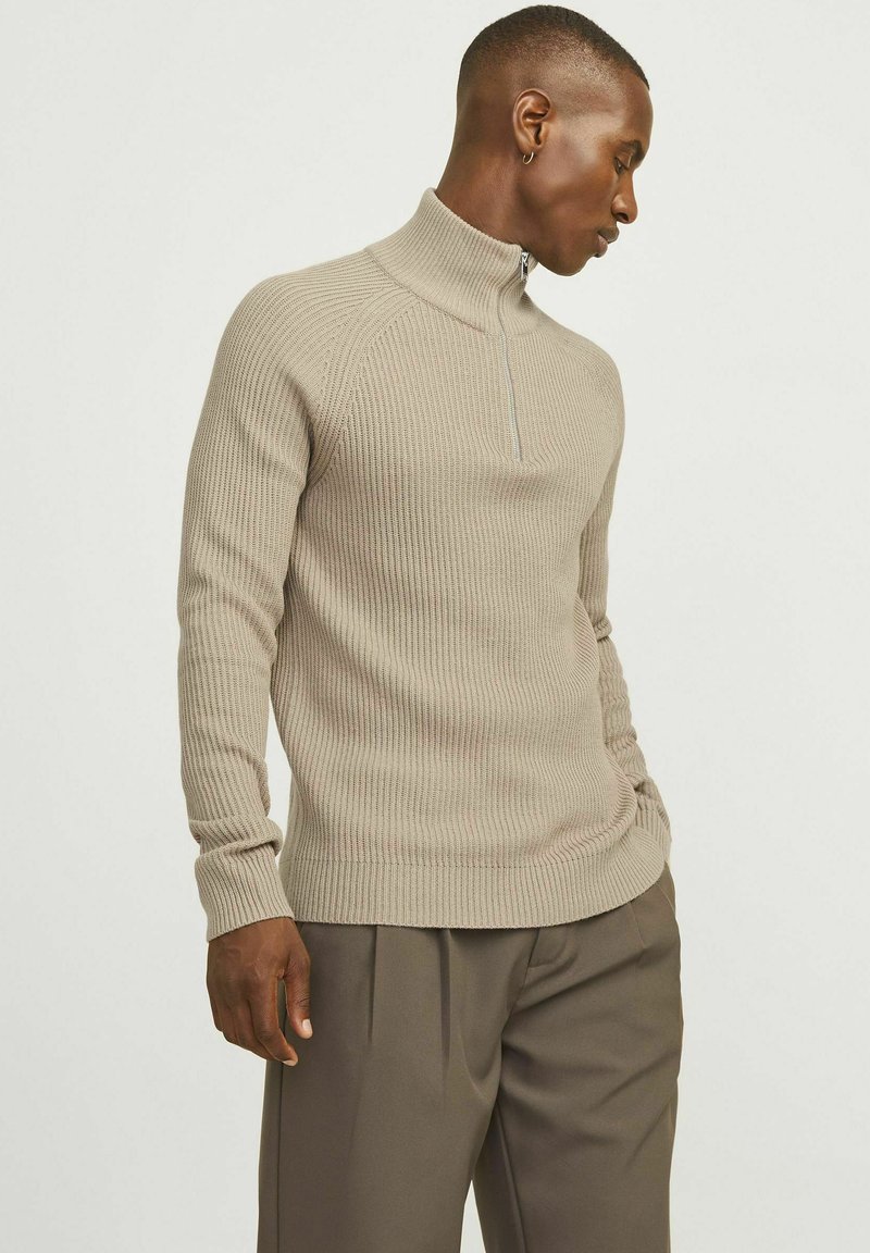 Beige geribbelde gebreide trui met een hoge kraag en een halve ritssluiting, voorzien van lange mouwen en een fitted ontwerp. Textuurdetails van de stof zijn zichtbaar.