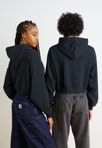Svart avkappede hettegensere med store hetter, båret med mørk denim og grå bukser. Sweatshirts har ribbede mansjetter og belter.