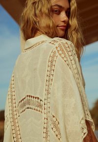 Kimono léger de couleur crème avec des détails et une bordure en dentelle intricate, présentant un design ouvert et une silhouette fluide.