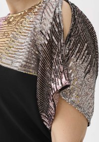 Detailní záběr na osobu v černých šatech s krátkými rukávy, pokrytých lesklými zlatými, stříbrnými a růžovými flitry.