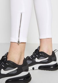 Leggings blancos con detalles de cremallera en el tobillo, combinados con zapatillas negras de Nike que presentan acentos grises y una suela de amortiguación prominente.