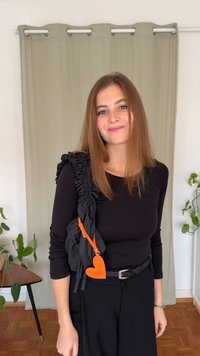 Črn zgornji del z dolgimi rokavi, kombiniran z visokim pasom črnih hlač. Črna torbica z volančki visi čez ramo, poudarjena z oranžno srčno oznako.