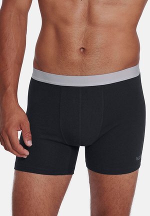 Mandligt torso iført sorte boksershorts med en lys grå linning, stående med venstre hånd ved siden mod en ensfarvet baggrund.