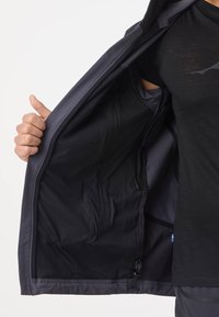 Persona che tiene aperta una giacca scura mostrando l'interno foderato in rete con una piccola tasca con zip, indossando una maglietta e pantaloni neri su sfondo bianco.