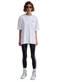 White oversized t-shirt with a blue logo, paired with black textured leggings featuring blue accents and white sneakers with blue details.