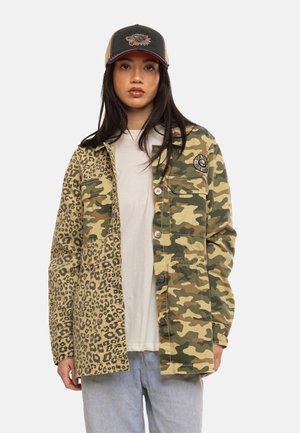 Jeune femme porte une veste moitié imprimé léopard, moitié camouflage sur une chemise blanche, associée à un jean bleu clair et une casquette beige et noire.