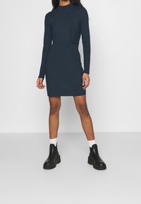 Marinblå långärmad klänning med en twistdetalj vid midjan, tillverkad av mjukt tyg. Kombinerad med kraftiga svarta stövlar och vita strumpor.