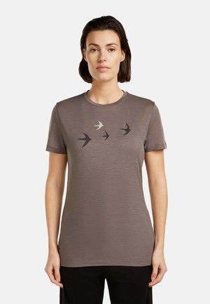 Frau mit kurzen dunklen Haaren, die ein taupefarbenes T-Shirt mit kurzen Ärmeln trägt, auf dessen Vorderseite vier fliegende Schwalbensilhouetten abgebildet sind.