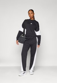 Conjunto atlético em preto e branco, composto por um sweatshirt de gola redonda e calças ajustadas. O material é macio, com uma textura suave e o logo da Adidas. Detalhes brancos destacam os lados.