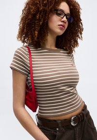 Νέα γυναίκα με σγουρά μαλλιά που φοράει μαύρα γυαλιά, μια ριγέ μπλούζα σε μπεζ και λευκό, σκούρο παντελόνι με ασημένια αγκράφα ζώνης και κόκκινη τσάντα ώμου.