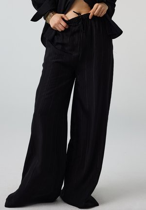 Personne ajustant un pantalon noir à jambes larges avec taille à cordon, vêtue d'une veste noire et de bracelets dorés sur fond clair uni.