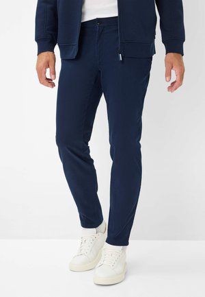 Navy blue slim-fit trousers made from a smooth fabric, featuring a button closure and pockets, paired with white sneakers.