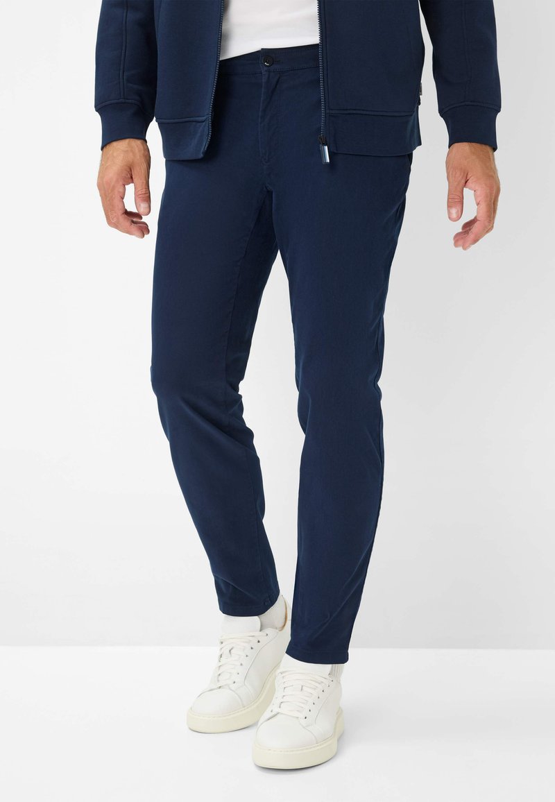 Marineblaue, taillierte Hose aus einem glatten Stoff, mit Knopfverschluss und Taschen, kombiniert mit weißen Sneakers.