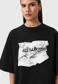 T-shirt nera oversize con grafica di carta accartocciata e testo "ALL SAINTS" con dettagli in più lingue. Collo rotondo e maniche corte.