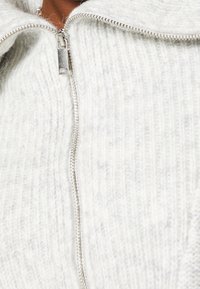 Lichtgrijze, gebreide sweater met ritssluiting en een gestructureerd patroon, voorzien van een zilveren rits en een zachte, pluizige uitstraling.