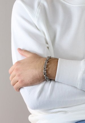 Silver metal chain bracelet worn on a hand, featuring interconnected links and a polished finish. The wrist is covered by a white textured sleeve.