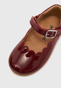 Γυαλιστερό σκ ουροκόκκινο παπούτσι Mary Jane με κυματιστές άκρες και ασημένιο λουράκι με πόρπη σε ανοιχτό φόντο.