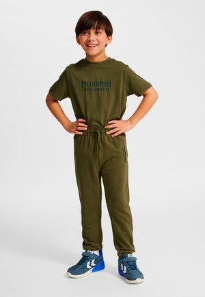 Grön bomullst-shirt och matchande joggers, med en logotyp på tröjan och byxorna. Barnet har på sig blå sportskor med vita detaljer.