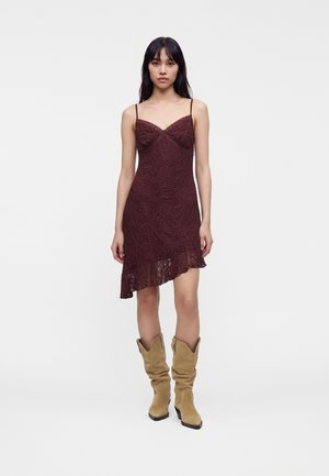 Femme portant une robe nuisette asymétrique en dentelle bordeaux à fines bretelles, associée à des bottes de cowboy souples en daim beige.
