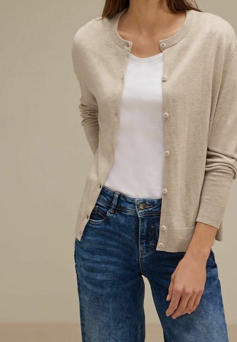 Femme portant un cardigan beige boutonné sur un t-shirt blanc et un jean bleu, debout avec une main posée près de sa hanche.