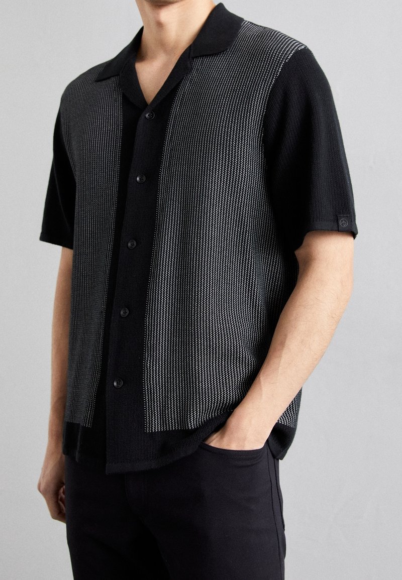 Homme portant une chemise noire à manches courtes avec des rayures grises texturées verticales à l'avant, associée à un pantalon noir.
