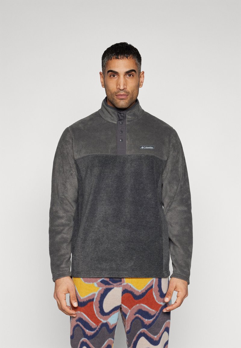 Grå fleece-pullover med ståkrave, der har en knaplukning og et logo-patch. Accentueret af farverige mønstrede bukser.