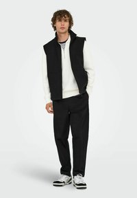 Schwarze ärmellose Weste über einem weißen Pullover, kombiniert mit schwarzen Hosen und schwarz-weißen Sneakers. Gestreiftes Hemd ist darunter sichtbar.