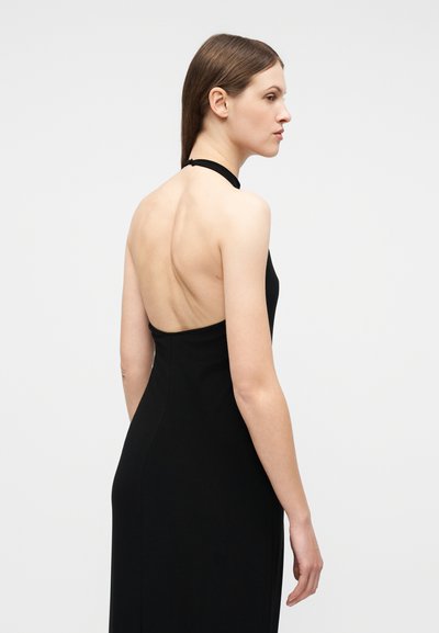 Γυναίκα με καστανά ίσια μαλλιά, φορώντας μαύρο αμάνικο φόρεμα με ανοιχτή πλάτη, κοιτάζοντας προς τα δεξιά σε απλό φόντο.