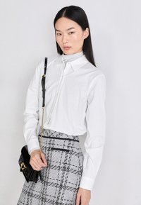 Chemise blanche à manches longues avec col et logo "HUGO" ; associée à une jupe grise à motifs et un petit sac noir avec une bandoulière chaîne.