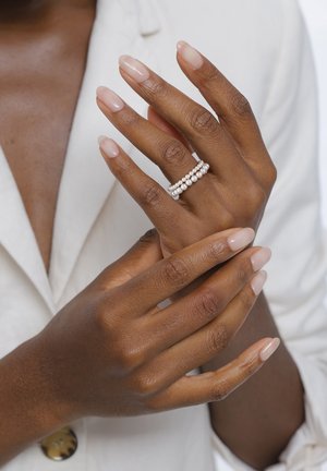 Mains à la peau foncée aux ongles naturels, dont une porte une bague à double rangée de perles, se touchant délicatement au-dessus d'une chemise blanche.