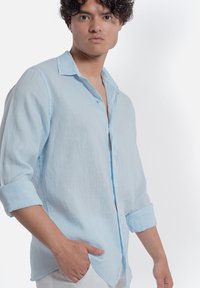 Camicia in lino azzurro chiaro con chiusura a bottoni sul davanti, polsini ripiegati e una vestibilità leggermente rilassata, realizzata in un tessuto testurizzato per garantire traspirabilità.