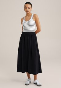 Gonna nera alla lunghezza del ginocchio con vita elastica, abbinata a una canotta bianca con righe orizzontali nere e scarpe sportive grigie.