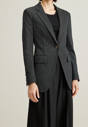 Femme portant un blazer à chevrons gris foncé à un seul bouton, sur un haut noir et un pantalon noir à jambes larges, devant un fond uni.