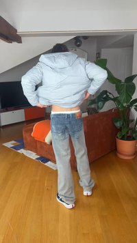 Egy szürke pufi dzsekis és farmernadrágos személy áll a nappaliban, háttal a nézőnek, kezét a csípőjén tartva igazítja a dzsekit egy narancssárga kanapé és egy nagy növény közelében.