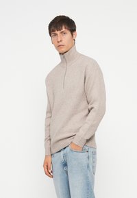 Suéter de punto beige con un cuello alto y cierre de media cremallera, con puños y dobladillo de canalé, combinado con jeans de mezclilla azul claro.