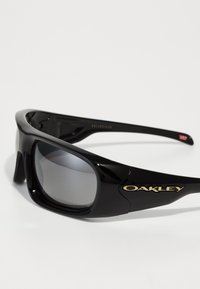 Óculos de sol pretos Oakley com lentes retangulares grandes e logótipo dourado na haste, mostrados sobre um fundo branco.