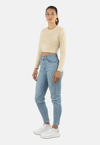 Beige Langarm-Cropped-Top, hellblaue High-Waist-Jeans, weiße Sneakers mit grauen Akzenten, stehend vor einem weißen Hintergrund.