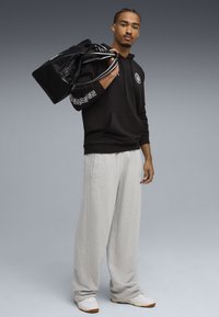 Mand iført sort hættetrøje og grå sweatpants, bærende sort duffelbag over skulderen, stående mod en grå baggrund.