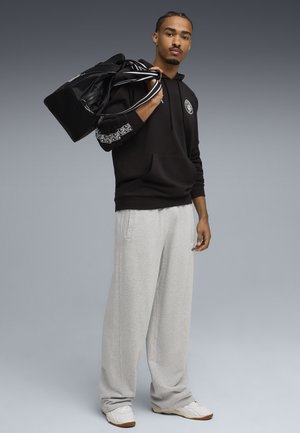 Mand iført sort hættetrøje og grå sweatpants, bærende sort duffelbag over skulderen, stående mod en grå baggrund.