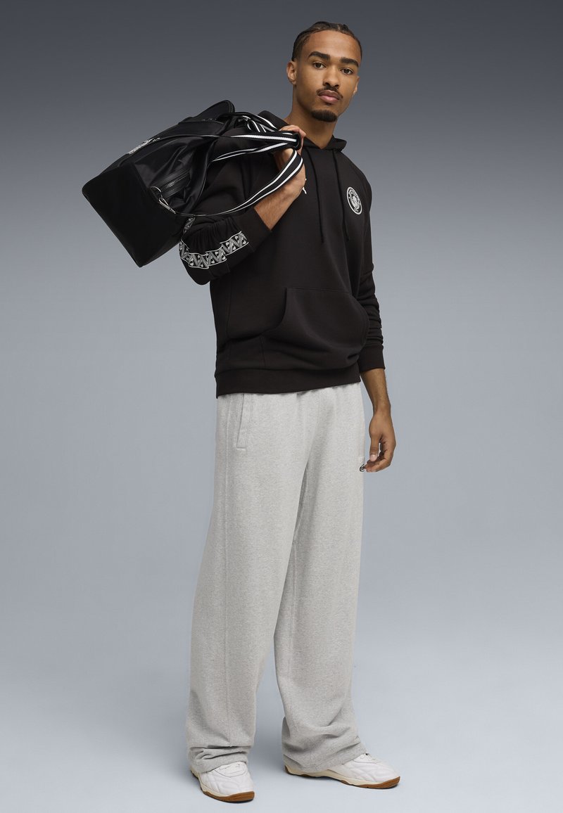 Mand iført sort hættetrøje og grå sweatpants, bærende sort duffelbag over skulderen, stående mod en grå baggrund.